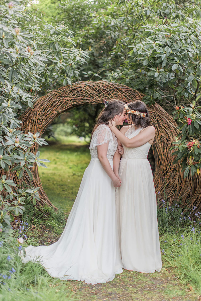 two brides wearing flower crowns standing kissing in a verdant and leafy wooded setting in front of a large woven wicker wood circular arch