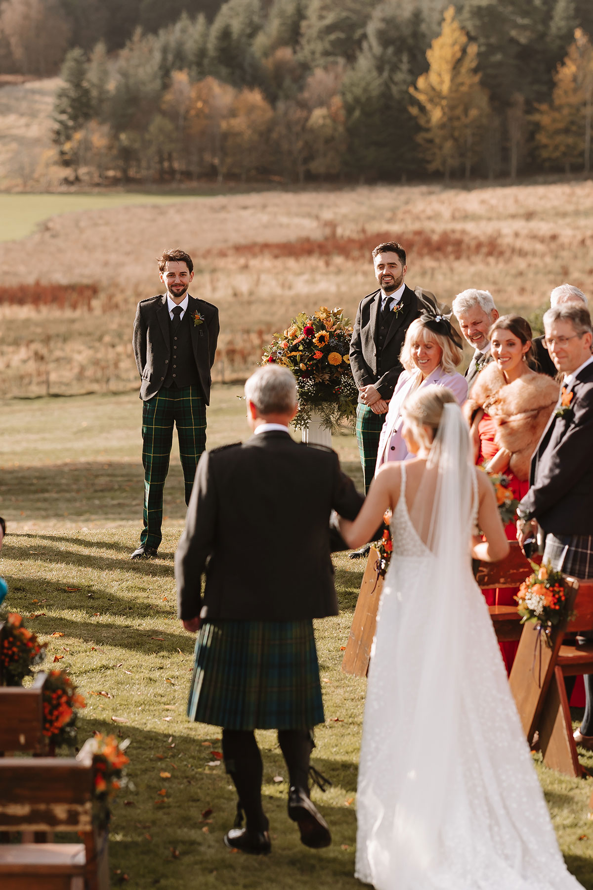 Bride in white dress walking down the aisle with her father towards the groom at outdoor ceremony.