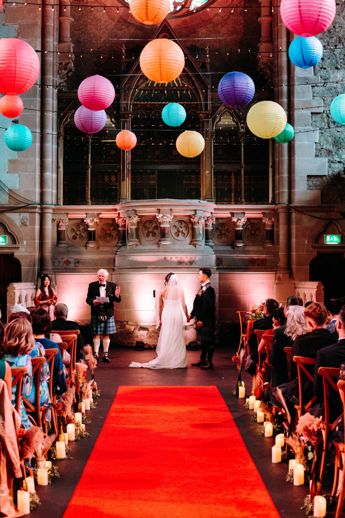 a bride and groom standing at the end of a red carpet in an ornate stone church style building with rows of seated wedding guests either side