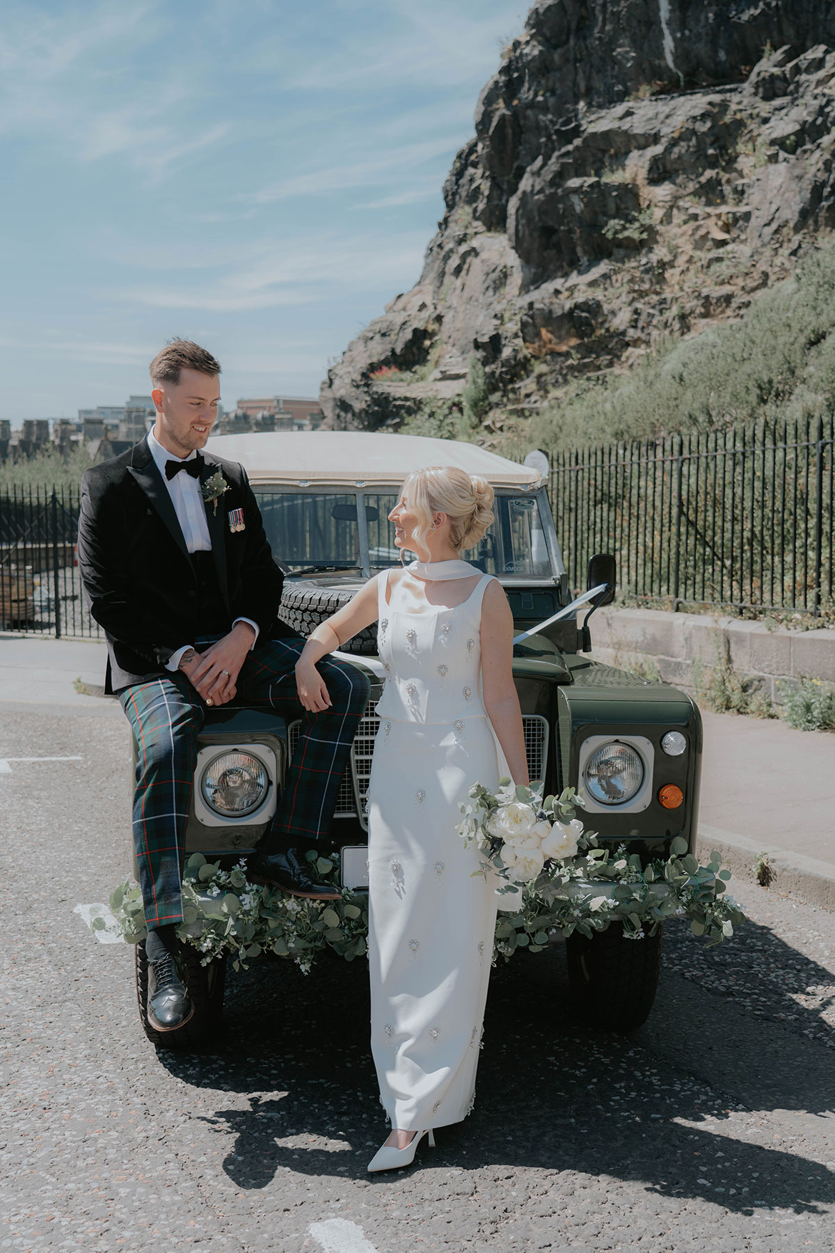 Newlyweds posing with a flower-decorated vintage Land Rover beside Edinburgh’s Salisbury Crags