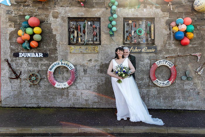 a windswept bride and groom standing in front of a stone building that's adorned with sea paraphernalia.