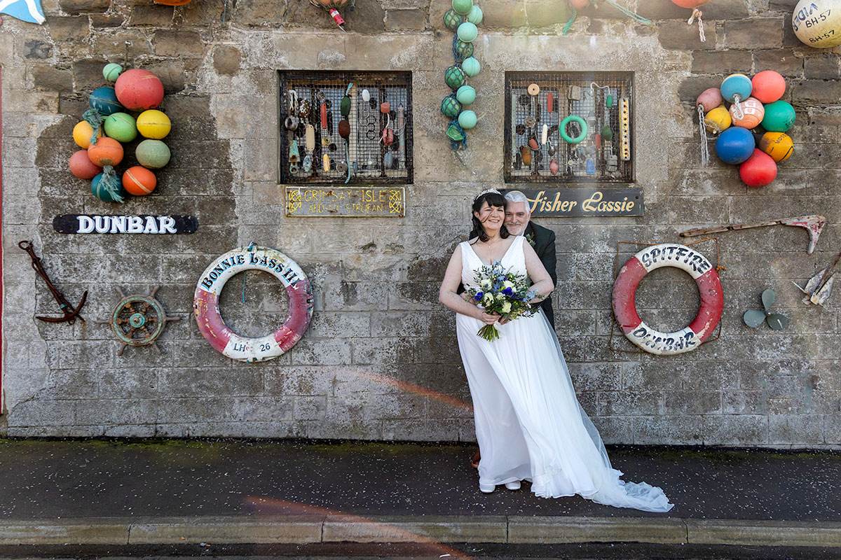 a windswept bride and groom standing in front of a stone building that's adorned with sea paraphernalia.