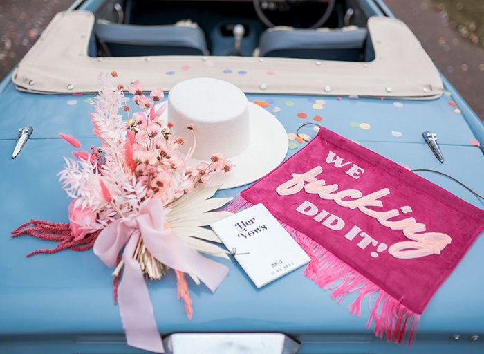 A white hat, a pink bouquet, a 'her vows' booklet and a pink 'we fucking did it' banner sit on top of the boot of a blue convertible