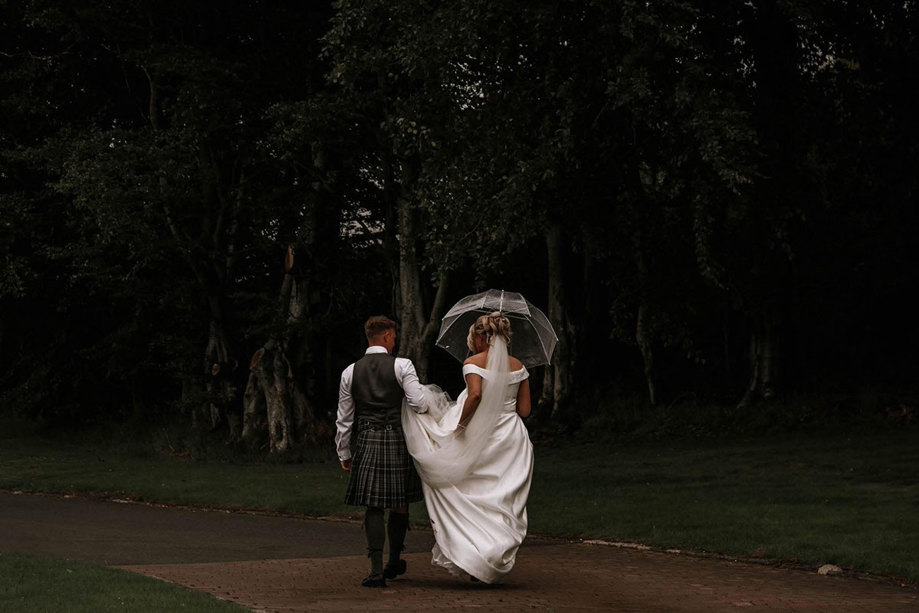 a bride and groom under an umbrella walking through woodland at The Vu countryside wedding venue central Scotland
