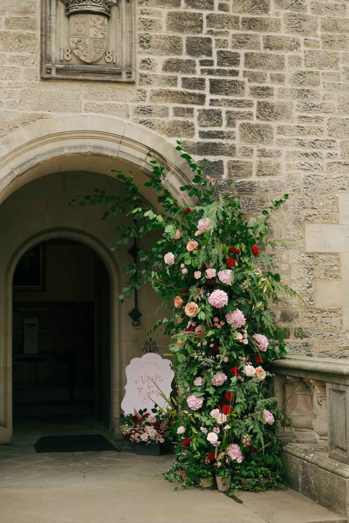 Large pink-and-red floral arrangement and personalised Nicole and Ryan wedding sign beside the stone entrance to Barnbougle Castle.