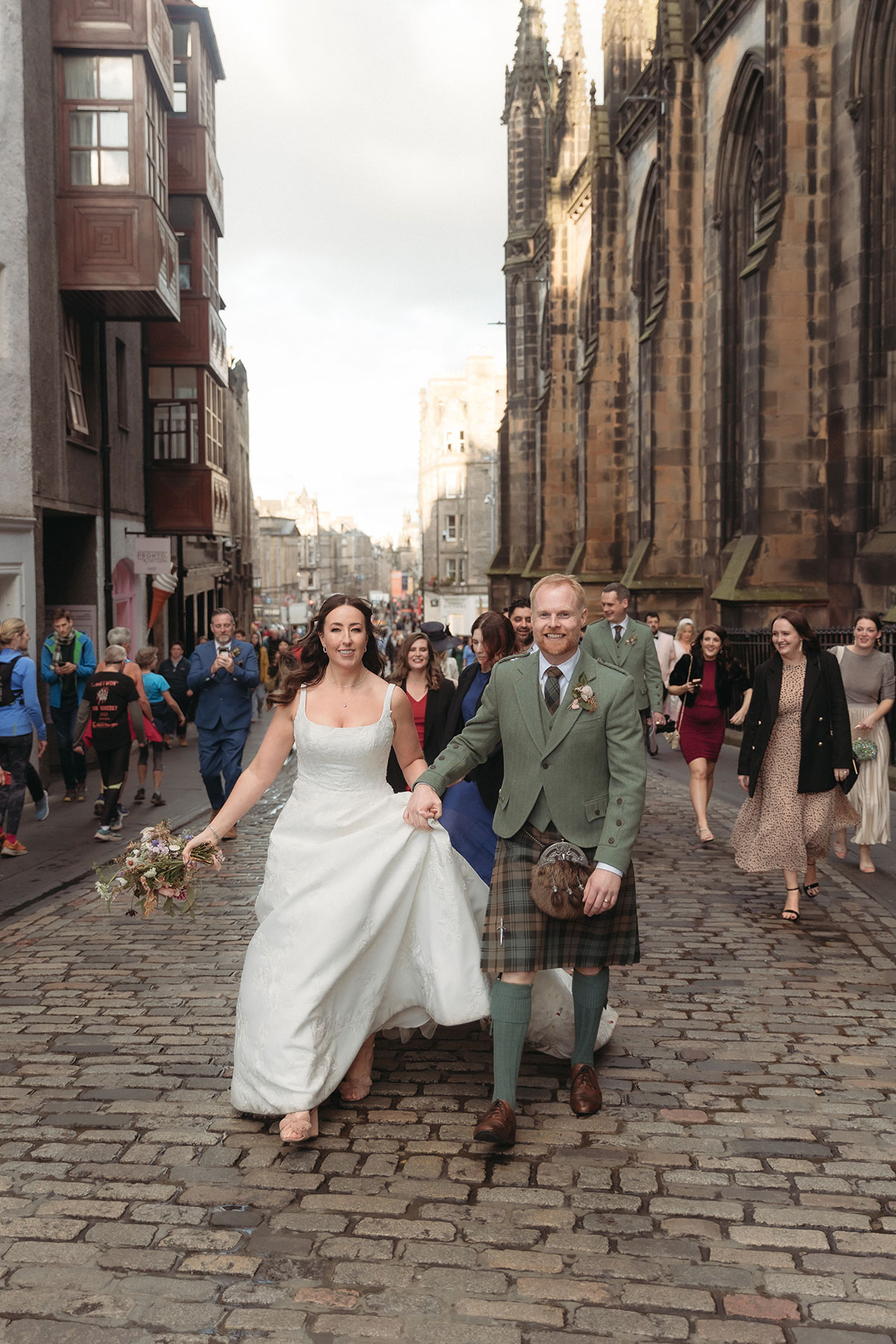 a smiling bride and groom walking along a cobbled street in Edinburgh with people behind them.