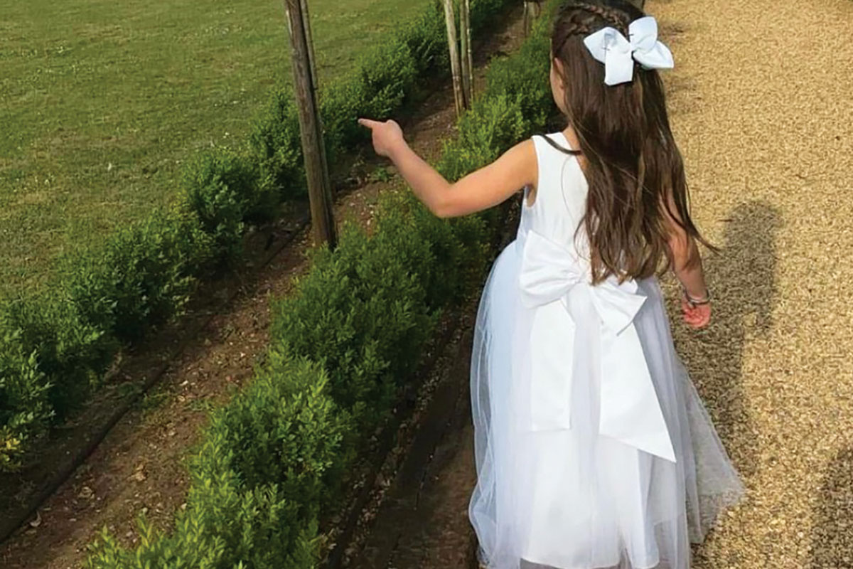 little girl in white dress with bow on the back and bow in her hair walks outside and points towards trees and bushes
