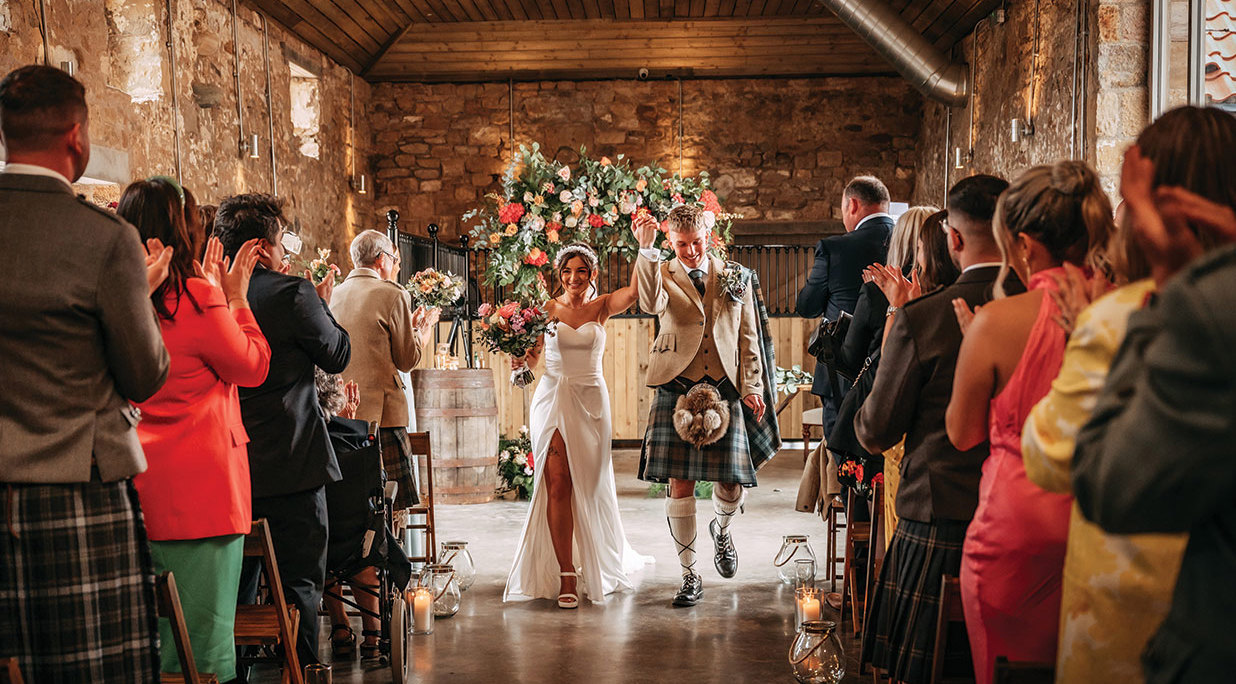 smiling bride and groom raise held hands aloft while walking on a concrete floor in a stone barn with wooden ceiling. There is a large flower display behind them.Rows of standing guests clap. 