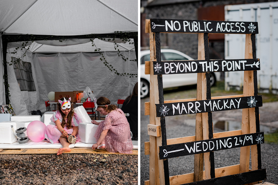 Two Children Wearing Pink Dresses And Fancy Dress Masks Sit On Floor Of White Marquee Building On Left And Black And White Painted Pallet Wedding Sign On Right