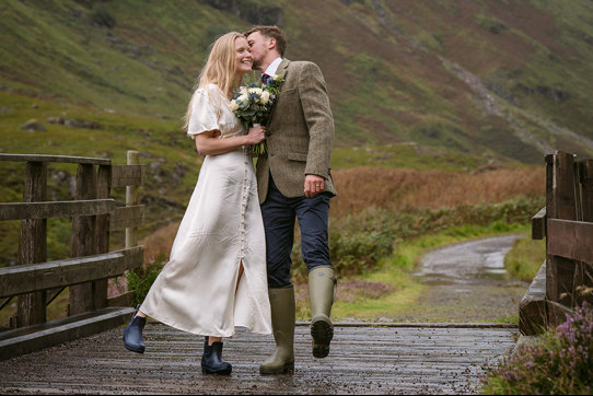 a groom kissing a bride on the cheek. They are both wearing wellies and standing on a wooden bridge in Glencoe
