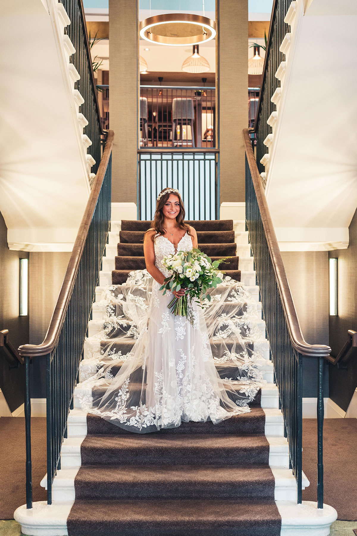 Bride standing on staircase at Clayton Hotel Glasgow with bouquet and flowing lace train