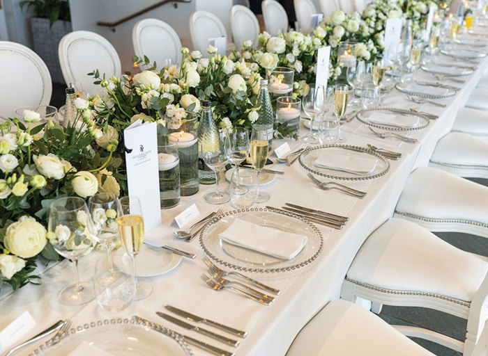 a long table with white high back chairs set for a dinner featuring clear charger plate with silver studs, abundant white flower decoration running down the middle and candles floating in glass holders.