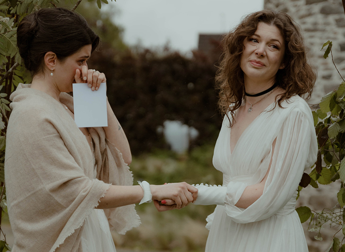 two emotional and tearful brides exchanging vowsoutside under a foliage-covered wire arch in a garden with old stone building in background