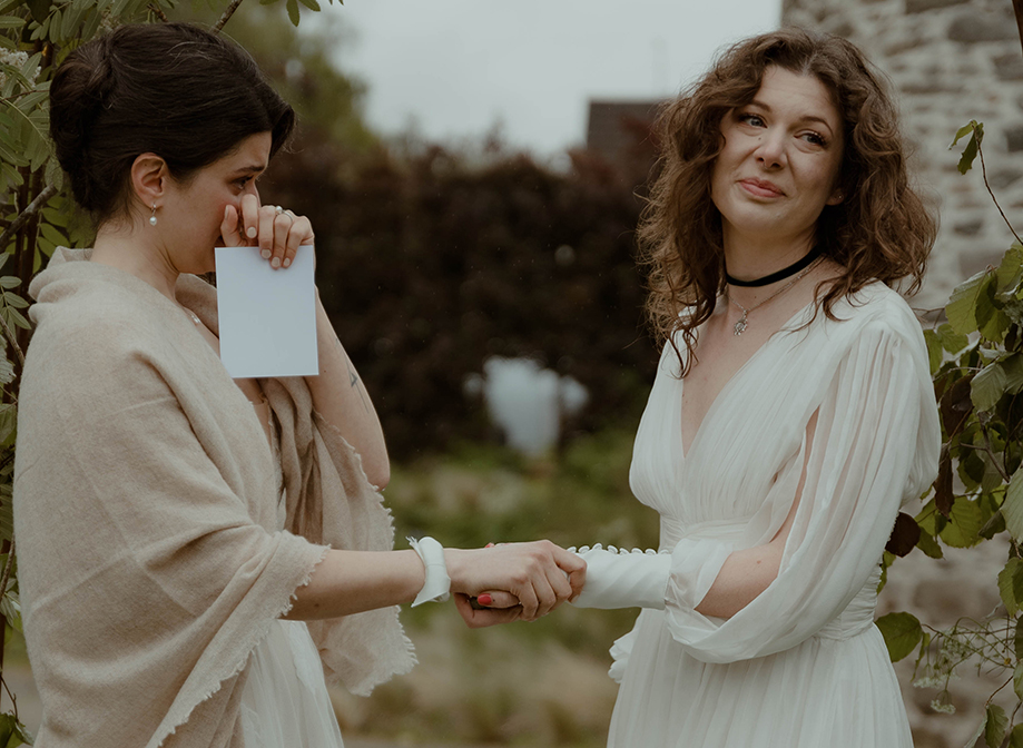 two emotional and tearful brides exchanging vowsoutside under a foliage-covered wire arch in a garden with old stone building in background
