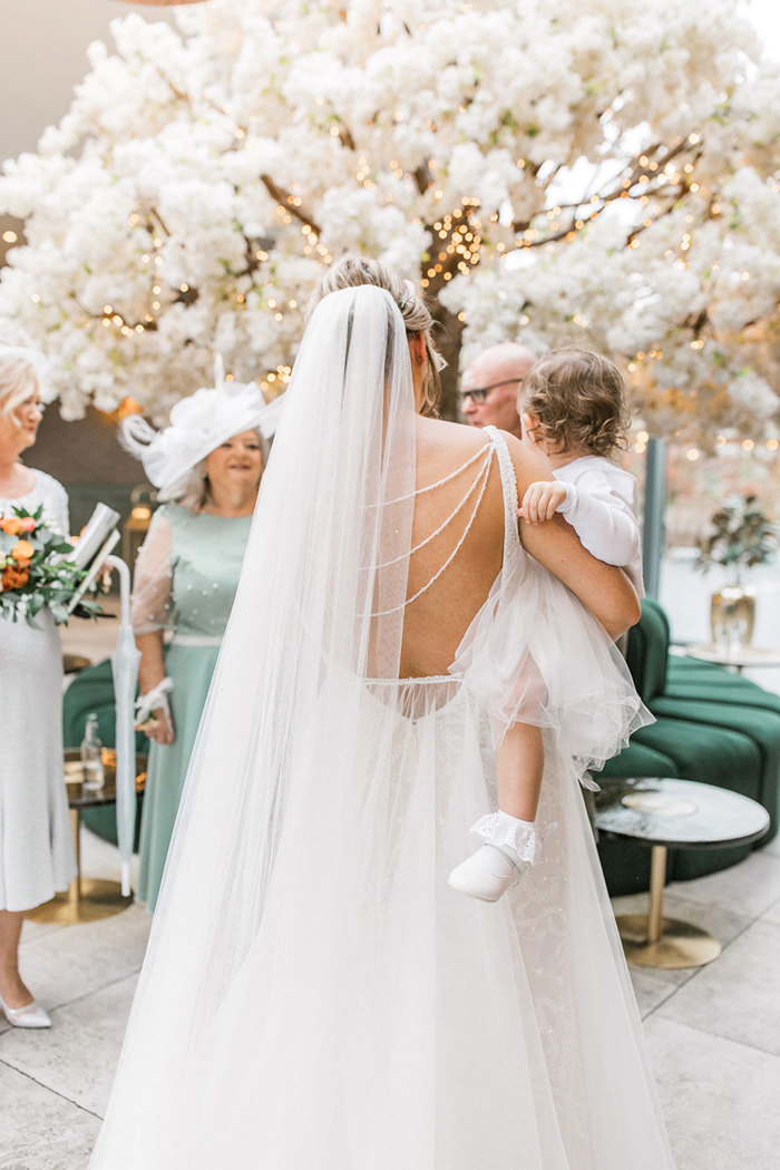 shot from the back of bride with low-back wedding gown with long veil, holding young girl in all-white outfit on her hip