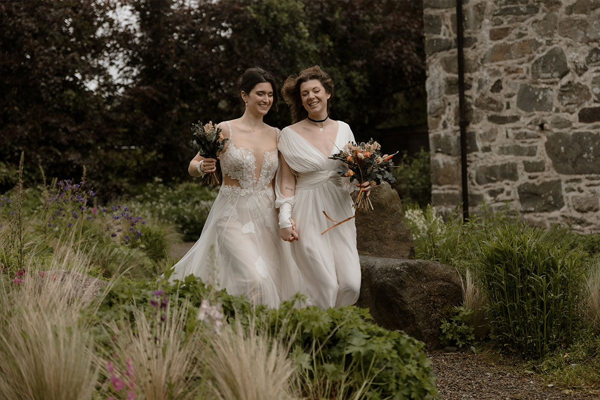 Two brides walk holding hands along a path in between rows of plants