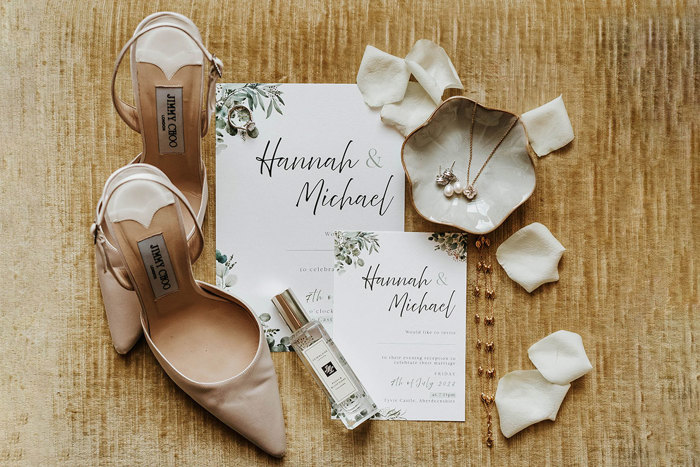 A pair of nude shoes, a perfume bottle, some white petals, a necklace, earrings and wedding invitations lying on a wooden surface 