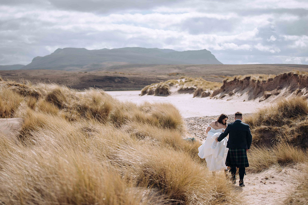 Bride lifts her dress as she and the groom walk through tall marram grass and sand dunes at Achnahaird Bay.