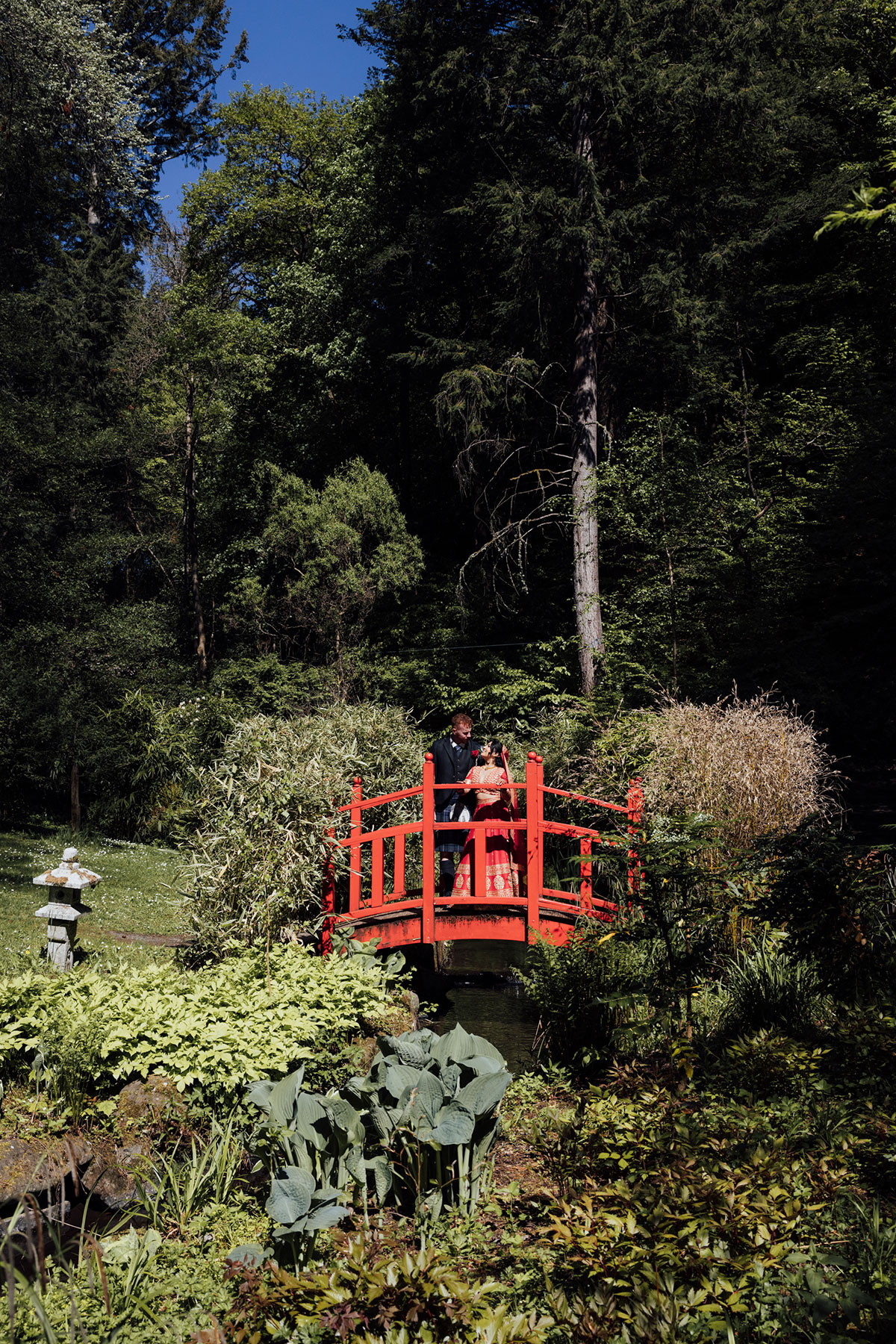Bride and groom standing on a red Japanese bridge in the gardens of Atholl Palace Hotel during their Scottish and Indian wedding portraits