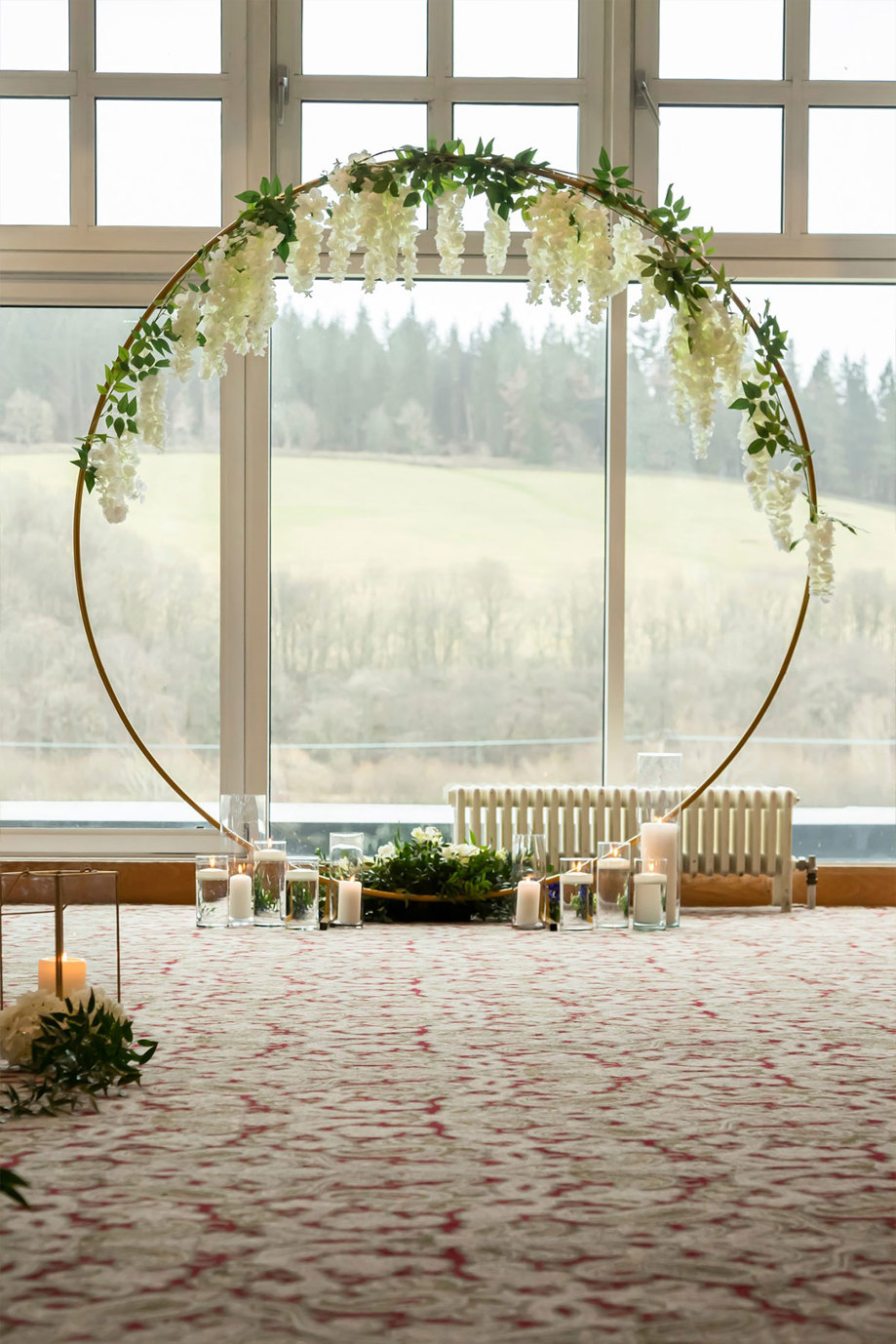 A moongate arch with foliage and white flowers hanging down around the hoop with candles in glass jars at the bottom set up in front of a window looking out to a forest