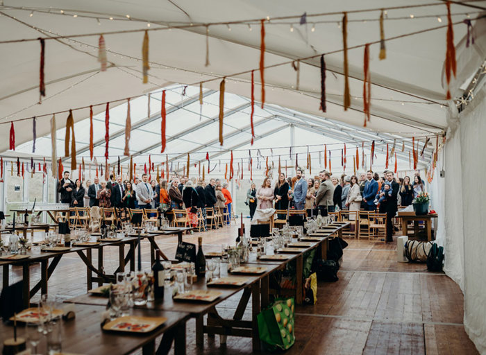 long tables in a marquee set for dinner with colourful paper streamers hanging from strings across the ceiling and a group of wedding guests at the back of the marquee