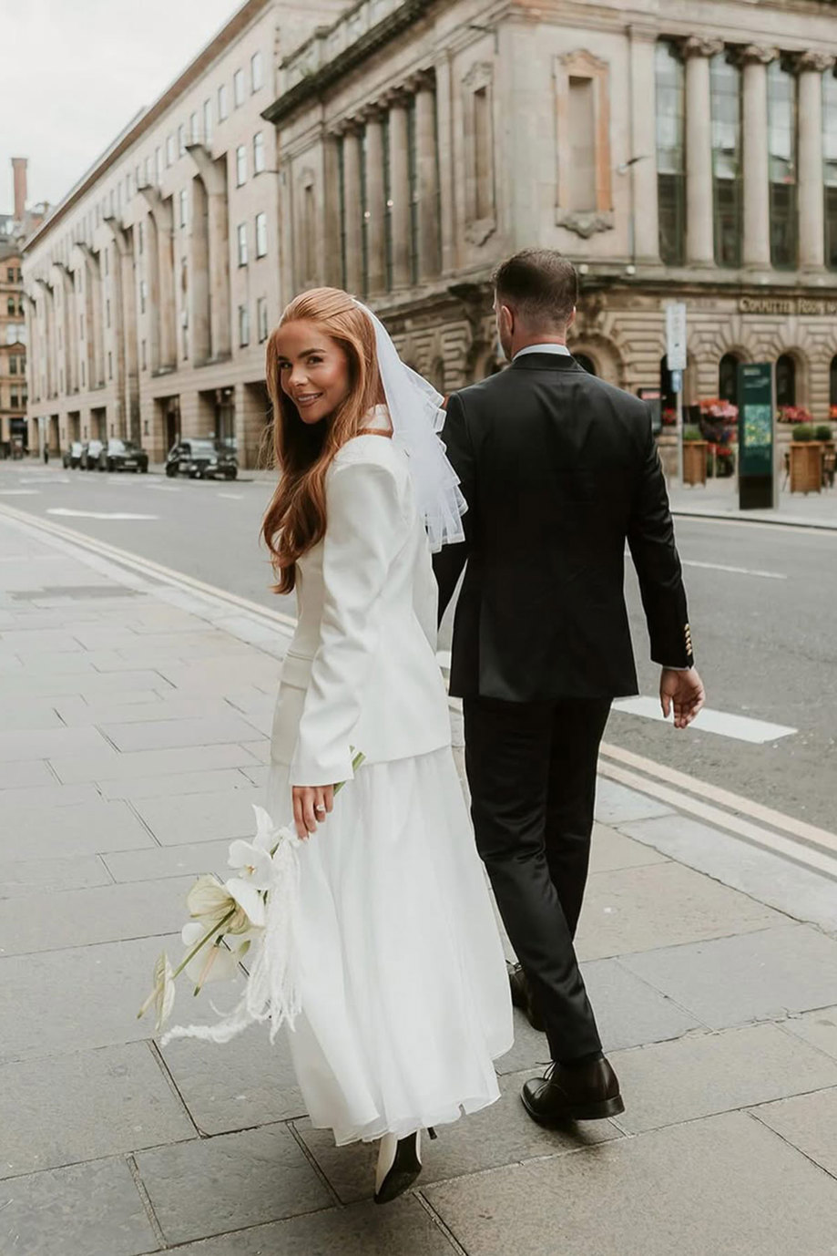 couple holding hands walking down city street, man in dark suit and red-haired woman in white jacket over a dress, a veil in her hair and lilies in her hand
