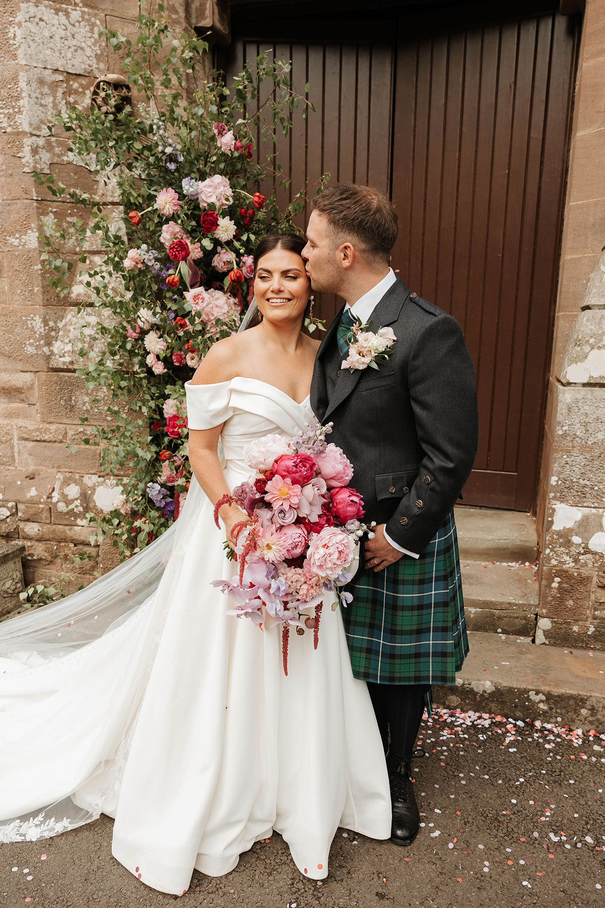 Groom kissing bride on forehead outside Drumtochty Castle Aberdeenshire with colourful wedding flowers
