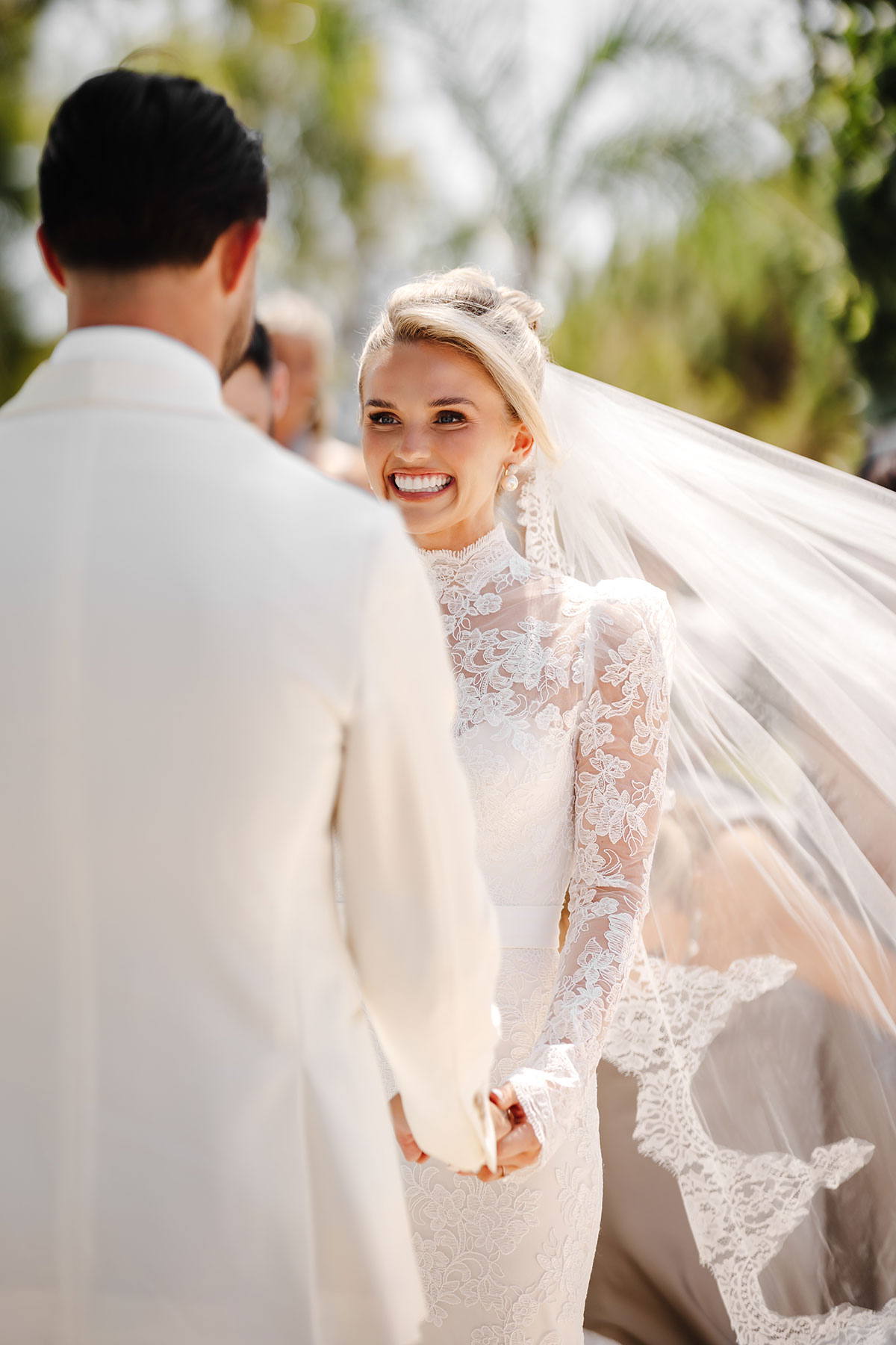 Bride in lace gown beams at groom as her veil catches the sunlight during the ceremony