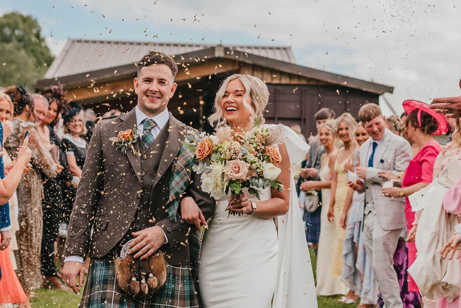 A couple walk arm in arm out of a barn as their guests throw confetti over them