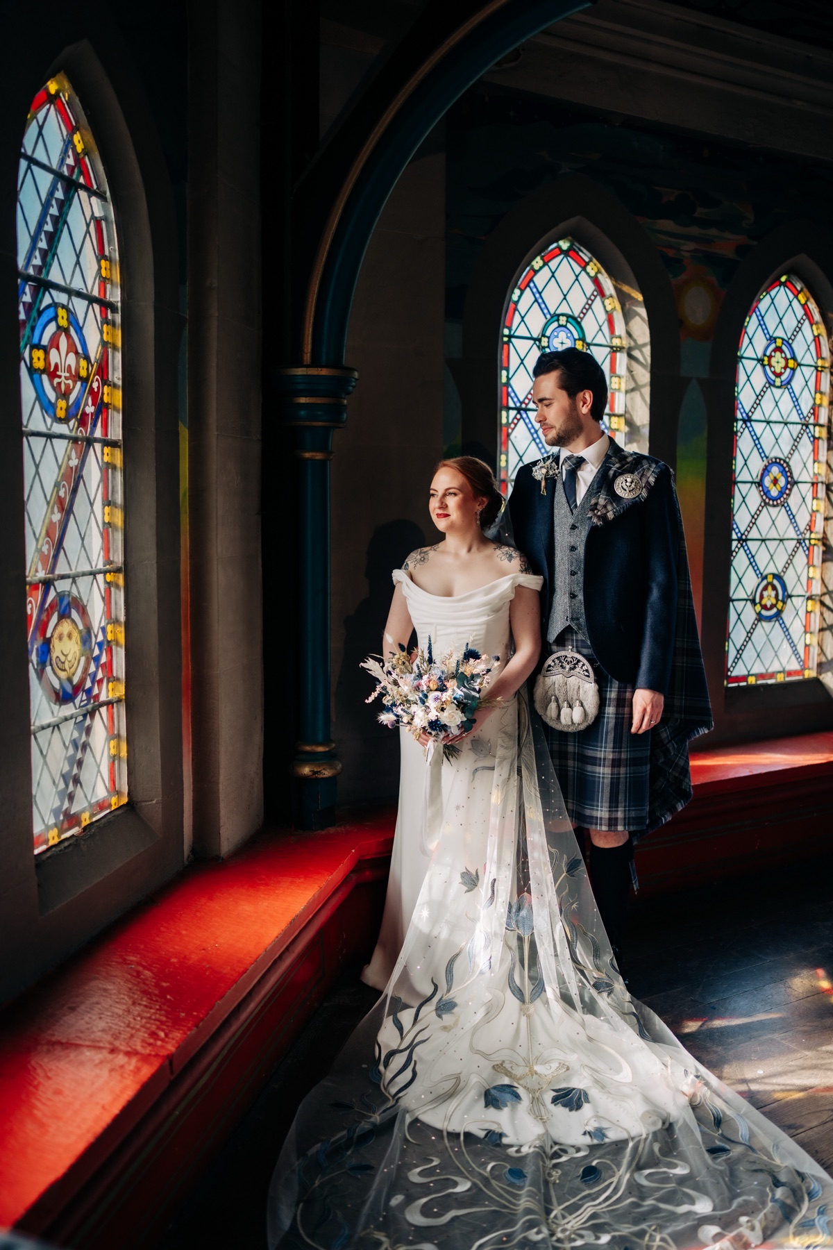 Robyn and James pose beside colourful stained-glass windows at Òran Mór in Glasgow, showing the blue floral embroidery on Robyn's wedding veil.