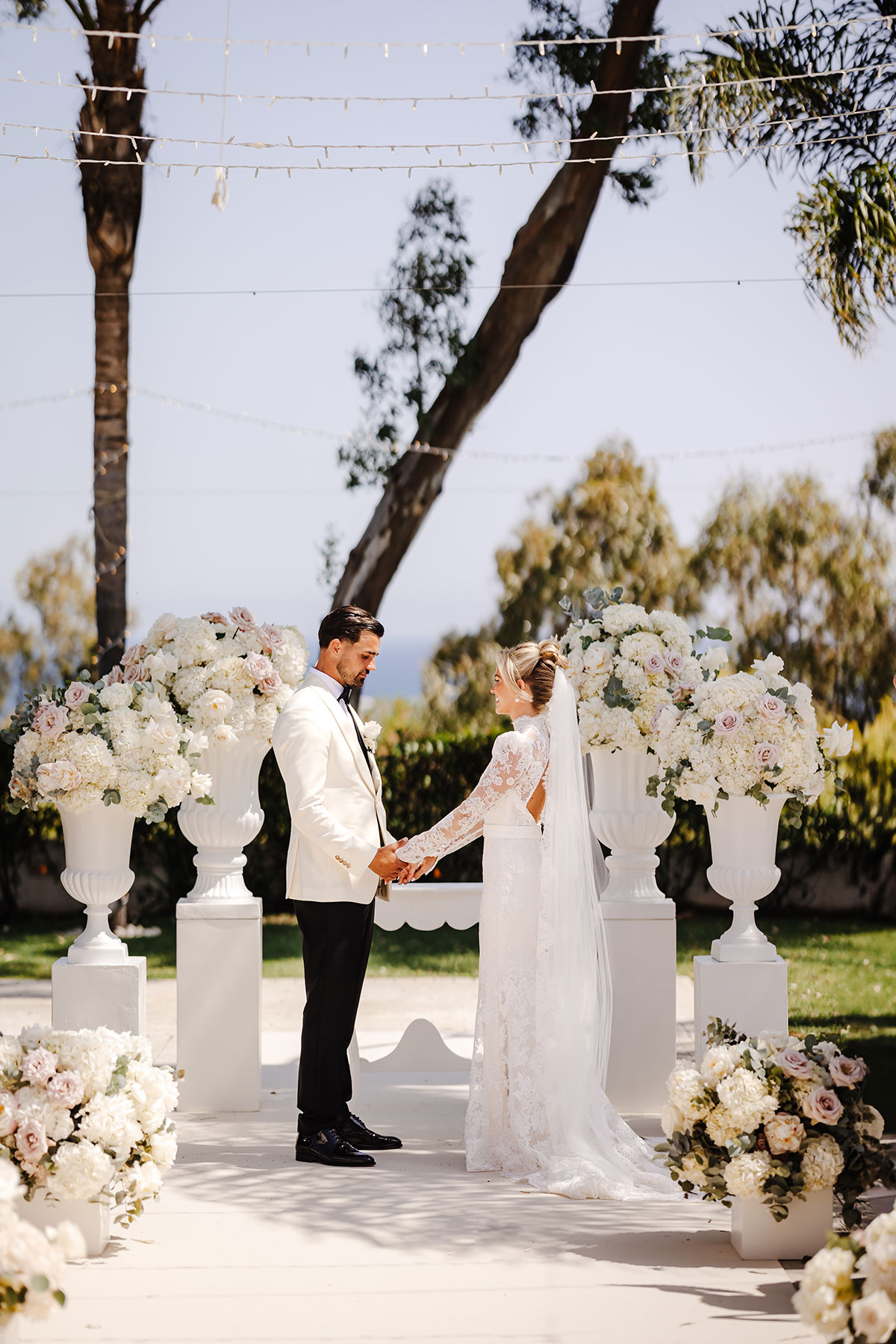 Couple stand hand in hand surrounded by tall white floral arrangements and trees