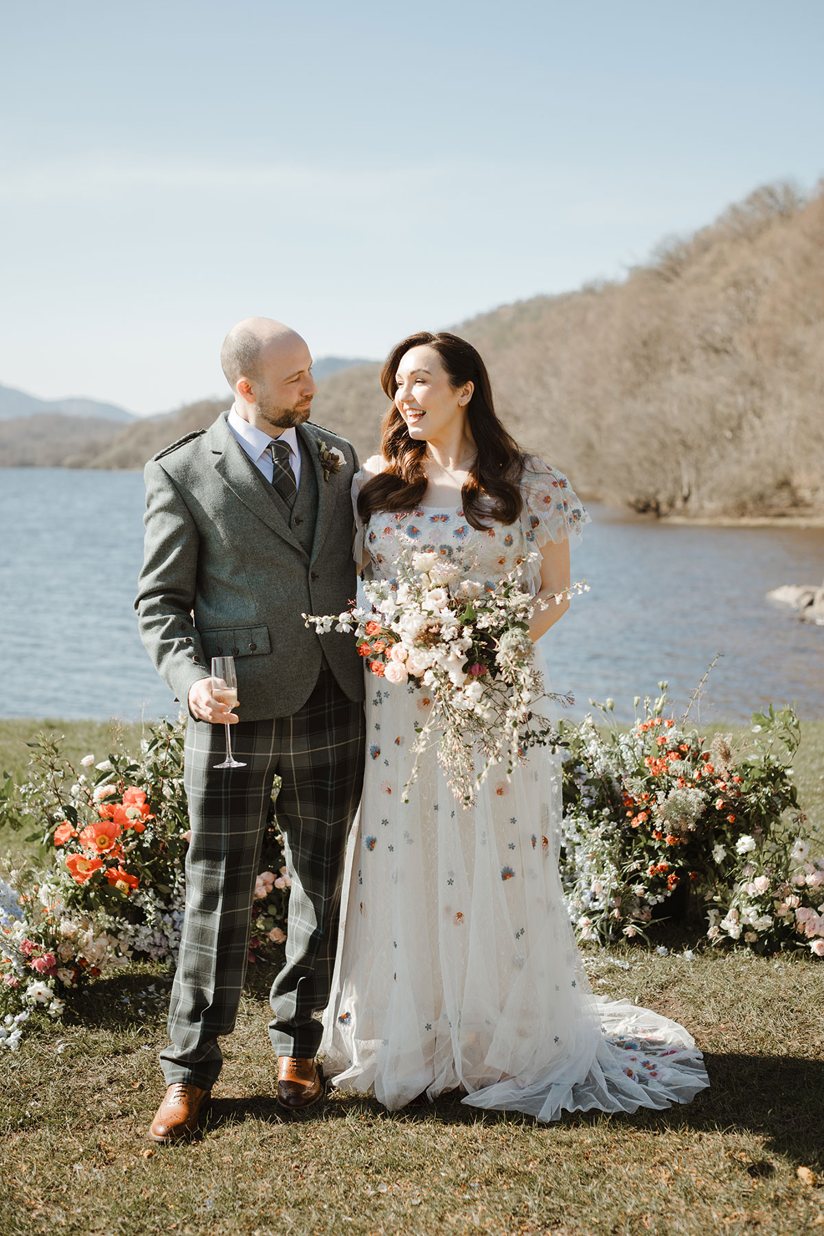 Bride and groom standing in front of the loch. Groom holds a glass of champaign, bride holds floral arrangement 