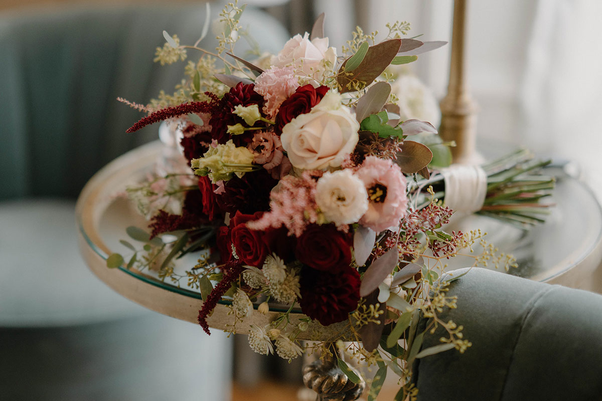 Autumn bridal bouquet with burgundy roses, blush blooms and dried florals on velvet chair