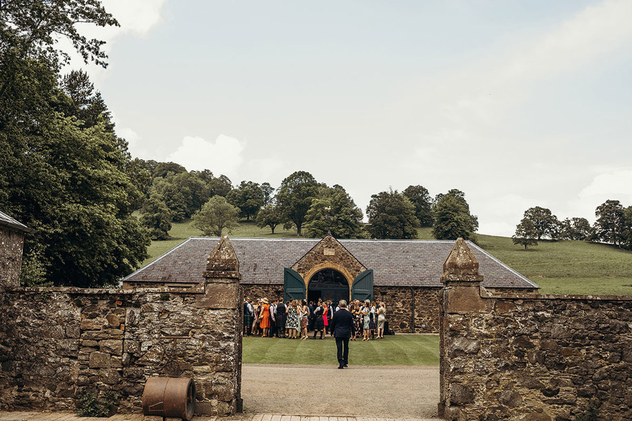 Wedding guests standing outside a stone byre with a hill and trees behind the venue