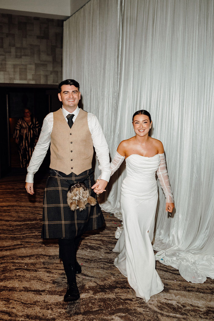 bride in strapless wedding dress with detached tulle sleeves holds hand of tall groom in beige and dark green kilt outfit indoors at a carpeted venue