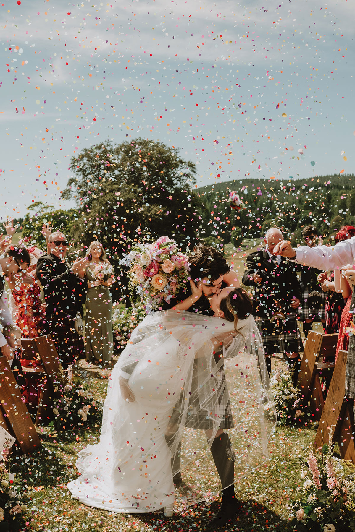 Bride and groom share a confetti kiss during an outdoor Scottish wedding ceremony surrounded by guests