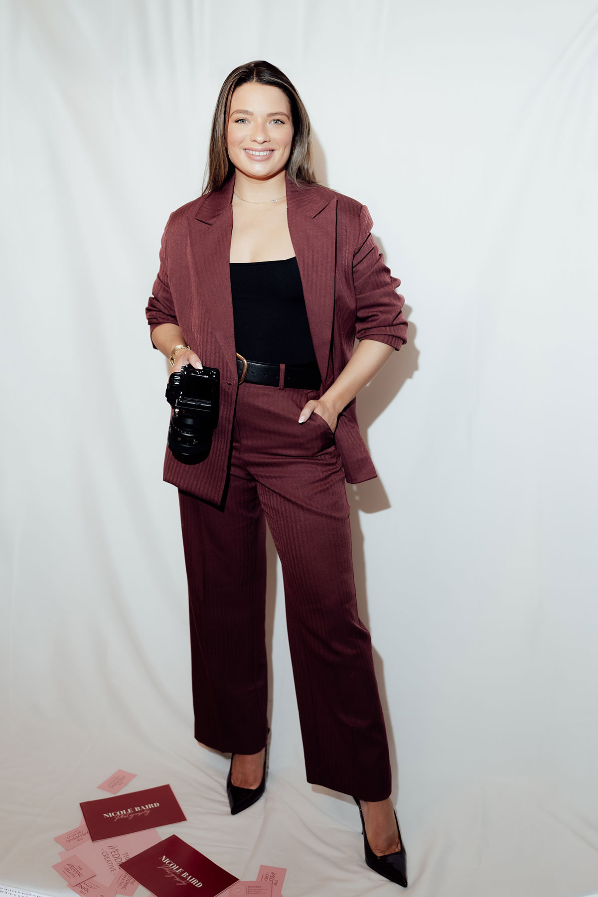 Wedding photographer posing with camera in burgundy suit at supplier showcase backdrop