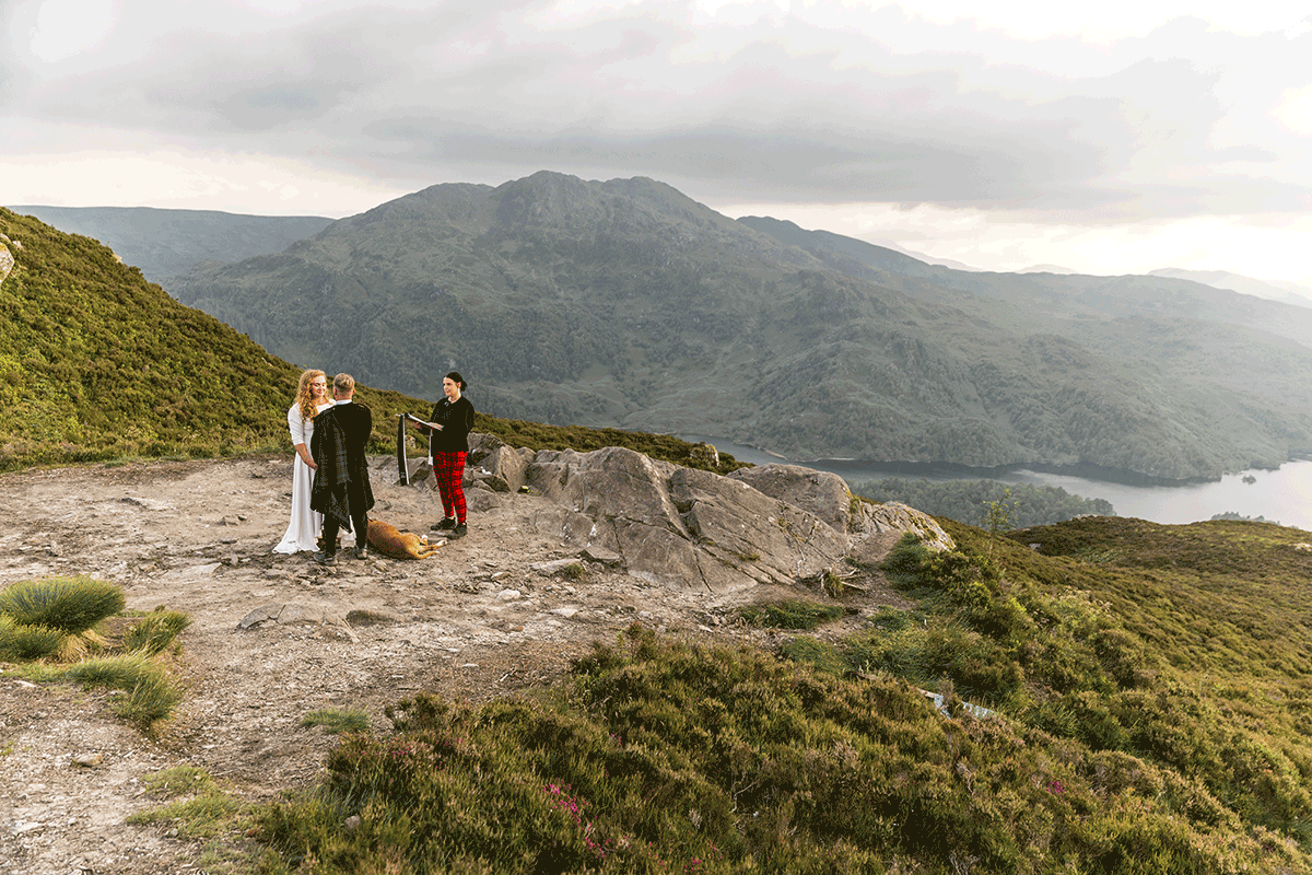 Wide landscape view of a Scottish mountain wedding ceremony overlooking a loch, with the couple, celebrant and dog surrounded by Highland scenery