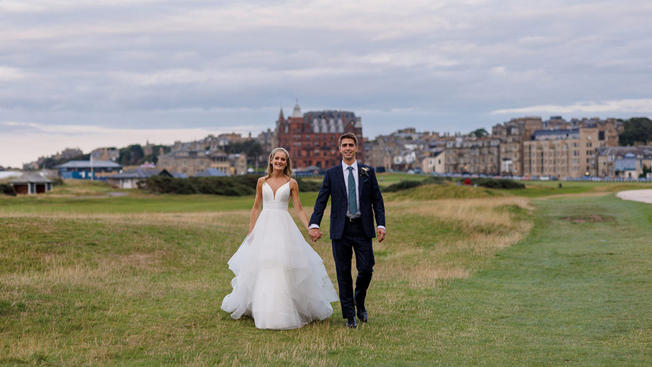 A joyful newlywed couple holding hands and walking across a grassy field in St Andrews with a picturesque cityscape in the background, captured by Duke Photography