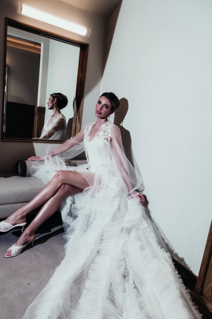 a bride sitting on a long pink bench wearing a ruffled and elaborate bridal robe from Eleganza Sposa next to a mirror