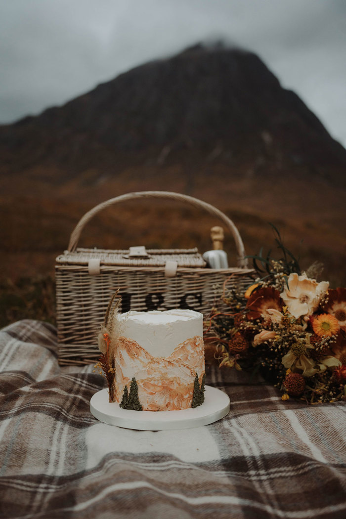 a wedding cake and picnic basket on a tartan blanket with Buachaille Etive Mòr in the background