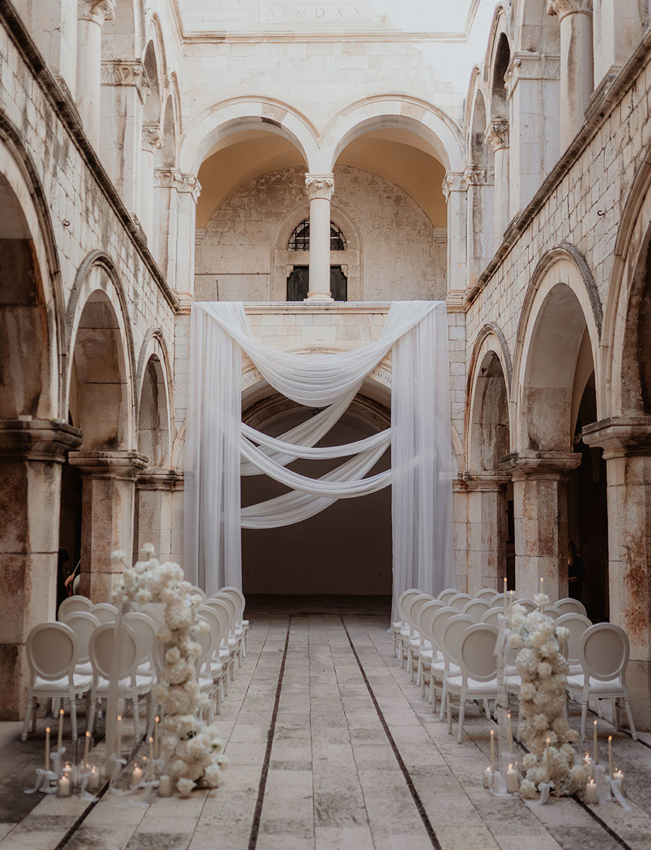 A stone courtyard set up for a wedding ceremony features white chairs, floral arrangements, and draped fabric under arched ceilings