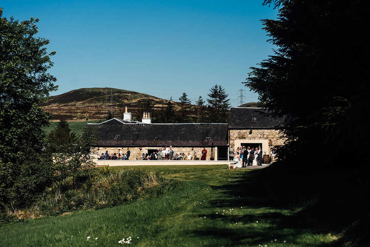 Guests gather outside the stone barn buildings at Blackshaw Barns in West Kilbride, surrounded by green countryside and hills under a clear blue sky.
