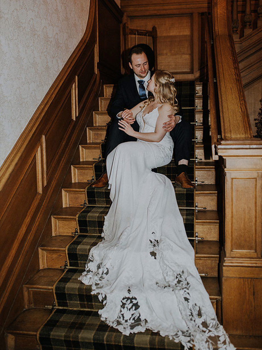 A bride and groom sit on a wooden staircase with a brown and green tartan runner with the bride's dress cascading down to the bottom step