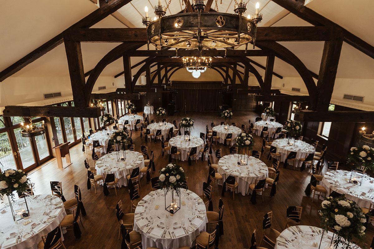 A large ballroom set for a wedding meal with round tables, tall floral centrepieces and exposed timber beams.