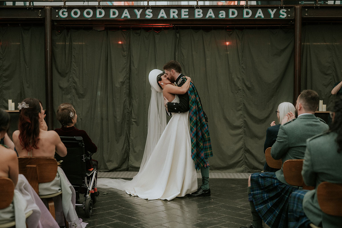 Bride and groom kiss during ceremony at Barras Art and Design Glasgow with guests seated and applauding