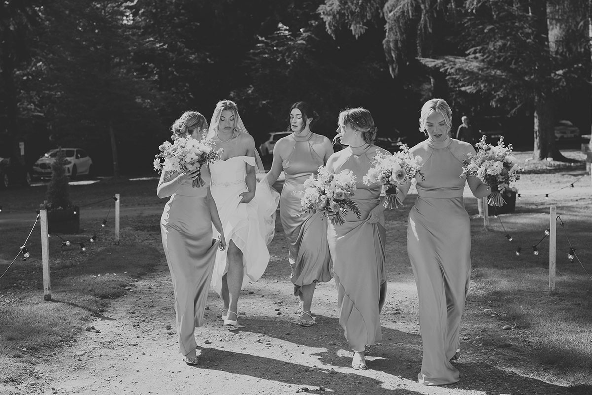 Black and white photo of bride and bridesmaids walking with bouquets along tree-lined path