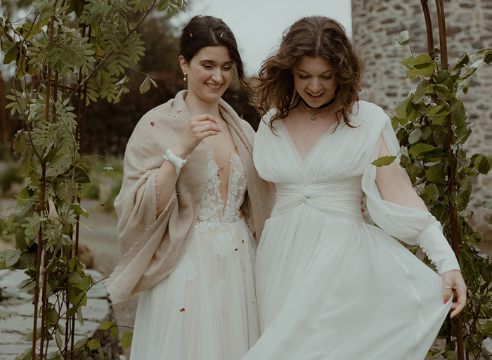 two joyful brides look downwards while standing outside under a foliage-covered wire arch in a garden with old stone building in background