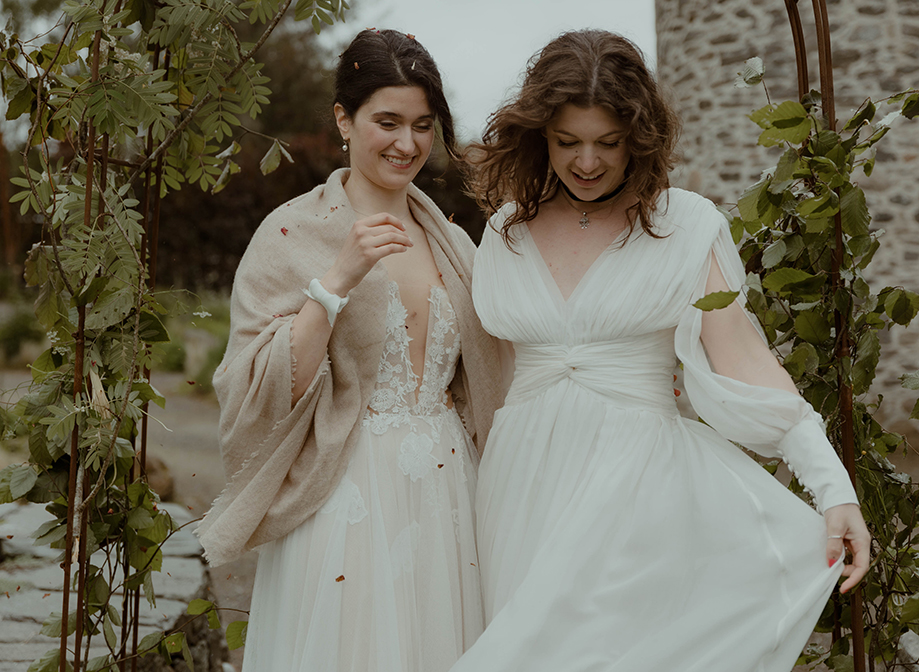 two joyful brides look downwards while standing outside under a foliage-covered wire arch in a garden with old stone building in background