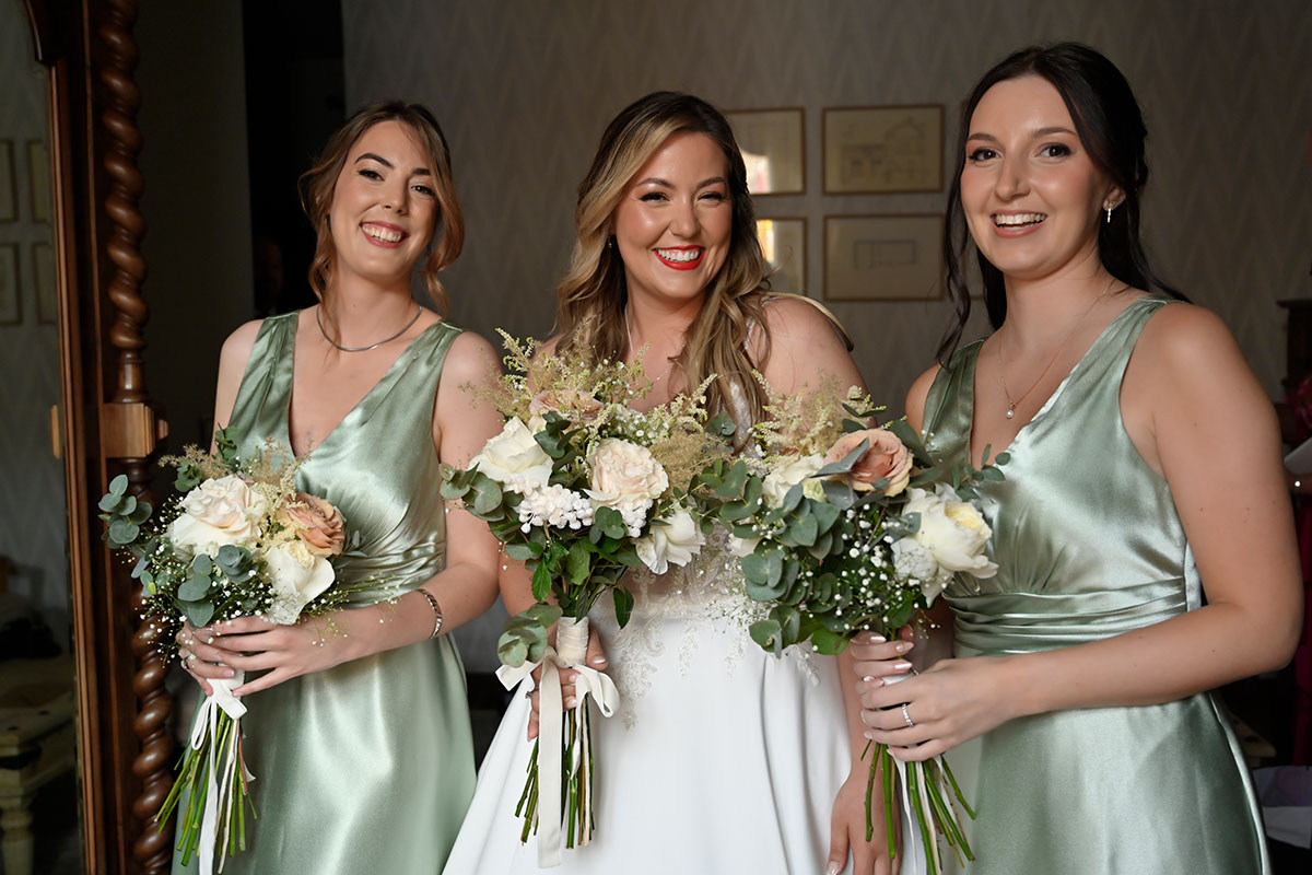 bride in white ballgown stands between two bridesmaids in shiny pale green dresses as they all smile and hold bouquets of neutral flowers